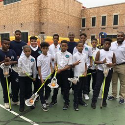 Students in white uniform shirts holding lacrosse sticks in a schoolyard
